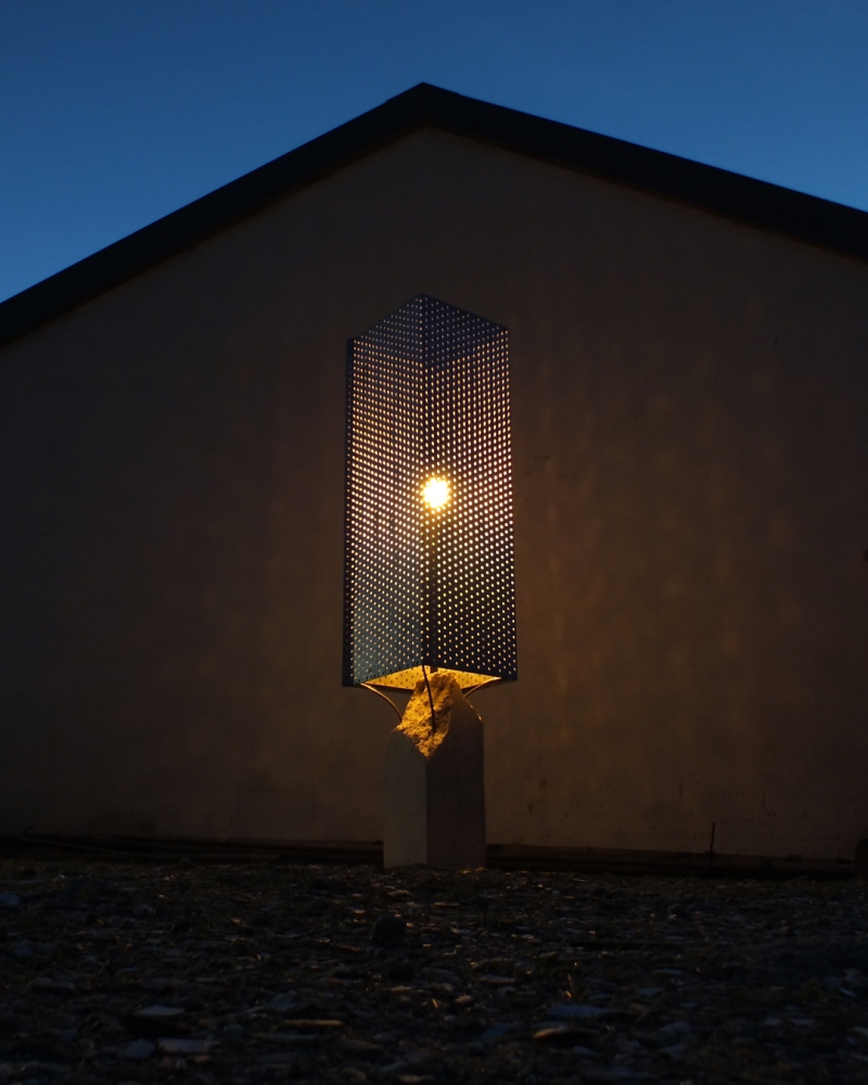 a nightime photo of a lamp made of concrete and perforated sheet steel